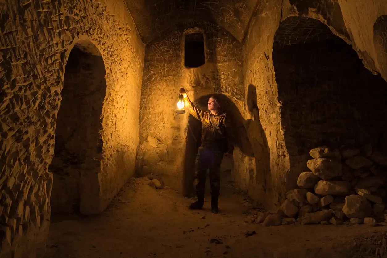Pessoa explorando uma caverna antiga à noite com lança de luz, em um ambiente rústico com paredes de pedra. Ideal para aventureiros e exploradores.