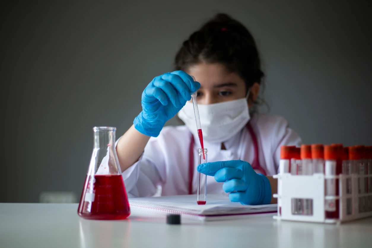 Jovem cientista realizando experimento com fluido vermelho em laboratório, usando jaleco e máscara de proteção facial. Ambiente profissional e higienizado.