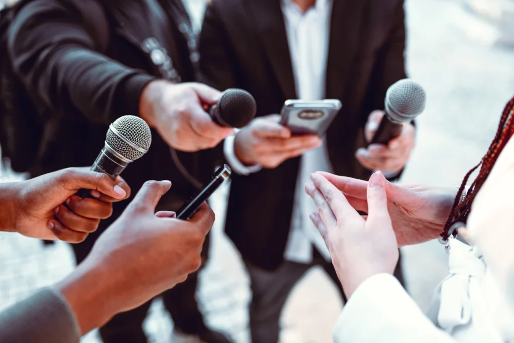Profissionais de imprensa realizando entrevista com uso de microfones, celulares e um tablet.