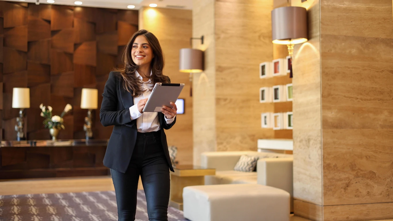 Mulher de negócios sorridente segurando um tablet em um ambiente moderno de escritório ou hotel, com decoração de madeira, almofadas e luminárias de parede.